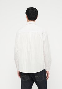 Άνδρας με την πλάτη στραμμένη φορώντας λευκό μακρυμάνικο πουκάμισο με λεπτές κάθετες ρίγες και σκούρο μπλε τζιν με κίτρινα ράμματα στις πίσω τσέπες.