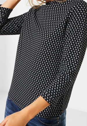 Kobieta w czarno-białej bluzce z długim rękawem o wzorze połączona z niebieskimi dżinsami, pozująca z uniesionym ramieniem przy głowie.