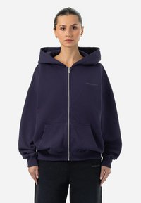 Námornícka modrá mikina na zips z mäkkej tkaniny s veľkou kapucňou, prednými vreckami a ribbed manžetami. Na hrudi sa nachádza reliéfne logo.