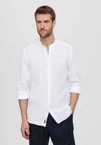 Camicia bianca di lino con colletto a banda, maniche lunghe ripiegate nei polsini e chiusura a bottoni. Presenta una texture sottile e un'etichetta laterale.