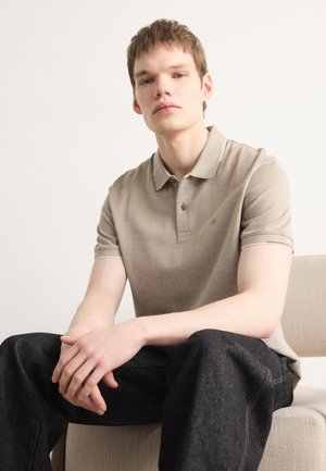Beige színű pólóing, gallérral, finom csíkokkal és gombos kihúzással, sötét farmernadrággal párosítva. Texturált anyag, minimalista márkázás.
