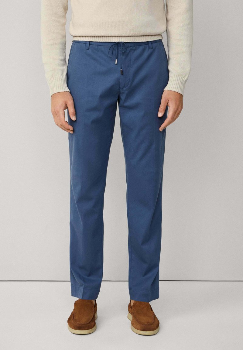 Pantalones azules hechos de una tela suave, con una cintura elástica y cordón ajustable, y piernas rectas. Combinados con calzado marrón.