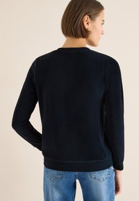 Laivastonsininen collegepaita, jossa on pitkät hihat, pyöreä pääntie, tekstuuri kangas, resorineulotut hihansuut ja helma. Päällä vaaleansiniset farkut.