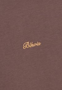 Brun stoff med en jevn tekstur som har "Blhvis" brodert med gyllen tråd nederst i høyre hjørne.