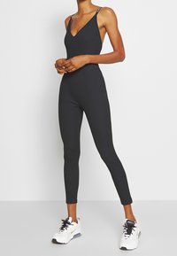 Kvinna står iförd en figursydd svart ärmlös jumpsuit och vita sportskor mot en helt vit bakgrund.