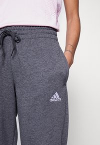 Pantalon de survêtement gris avec une taille élastique et cordon de serrage, poches avant et un logo Adidas blanc sur la cuisse gauche.