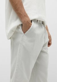Pantaloni in cotone grigio chiaro con cintura elastica, tasche laterali e dettagli di cucitura a vista. Indossati con una camicia bianca larga.