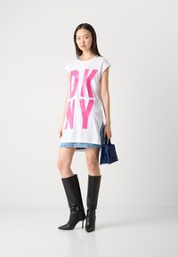 Abito t-shirt bianco oversize con grande stampa rosa "DKNY", abbinato a una mini gonna di jeans e stivali neri alti fino al ginocchio con tacco a spillo. Piccola borsa blu.