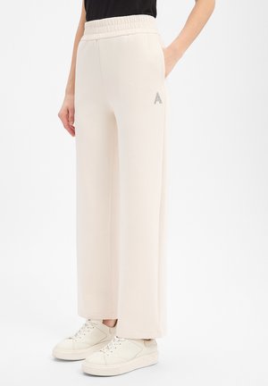 Person trägt cremefarbene, weit geschnittene Hose mit elastischem Bund und weiße Sneaker, steht mit einer Hand in der Tasche.