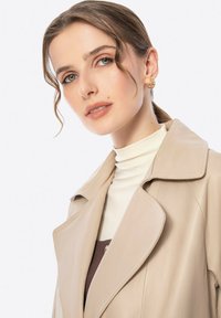 Cappotto trench in pelle beige con ampio colletto, indossato sopra una maglia bianca. Presenta una texture morbida e linee pulite, evidenziando un design moderno.