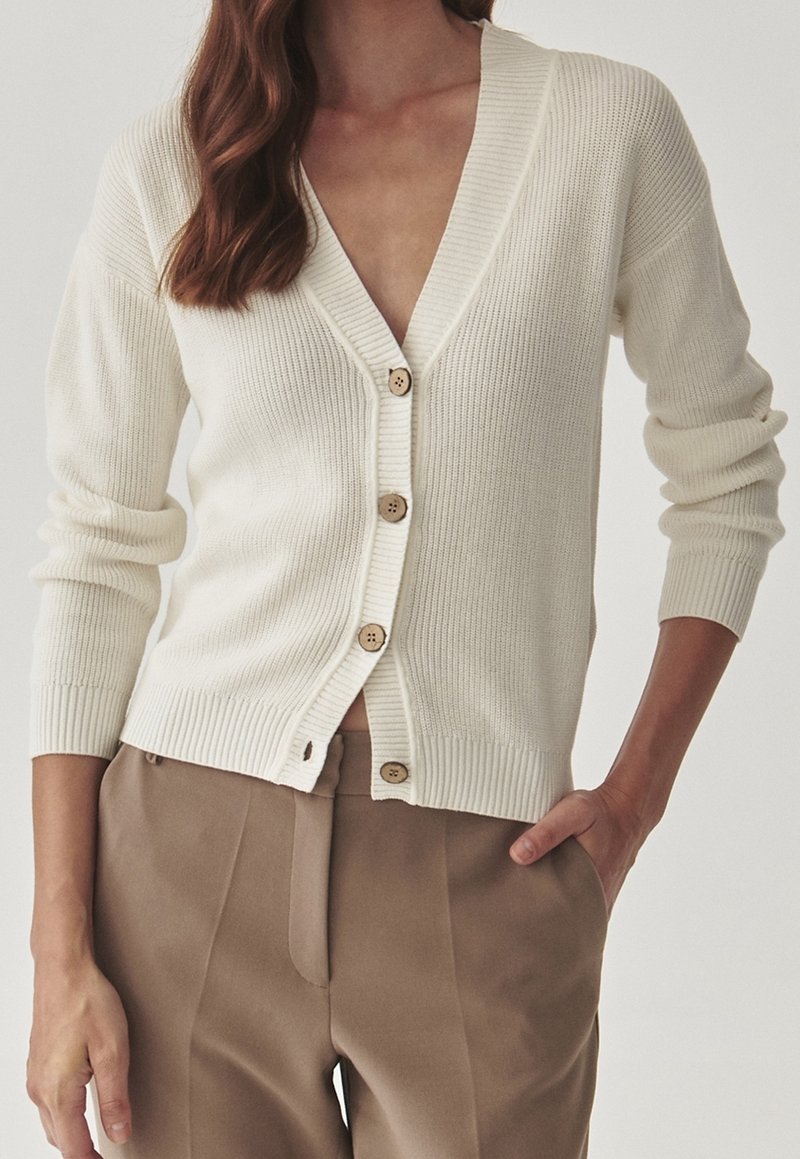 Cárdigan crema con un profundo escote en V, textura acanalada y cinco botones de madera en el frente. Mangas largas ajustadas y un corte a la cadera.