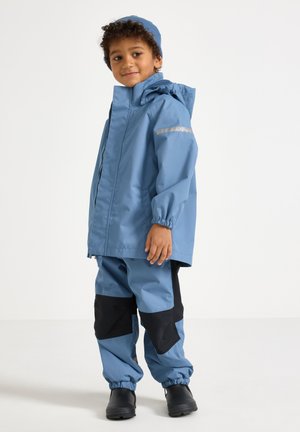 Lapsi, joka on pukeutunut siniseen vedenpitävään takkiin ja housuihin, joissa on mustat polvipaikat, mustiin saappaisiin ja siniseen neulepipoon, seisoo valkoisella taustalla.