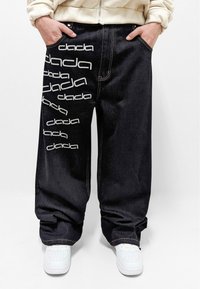 Pantalones de mezclilla negra con corte oversized. Presentan texto blanco "dada" y costuras visibles. Bolsillos frontales y dobladillos con puños, usados con zapatillas blancas.