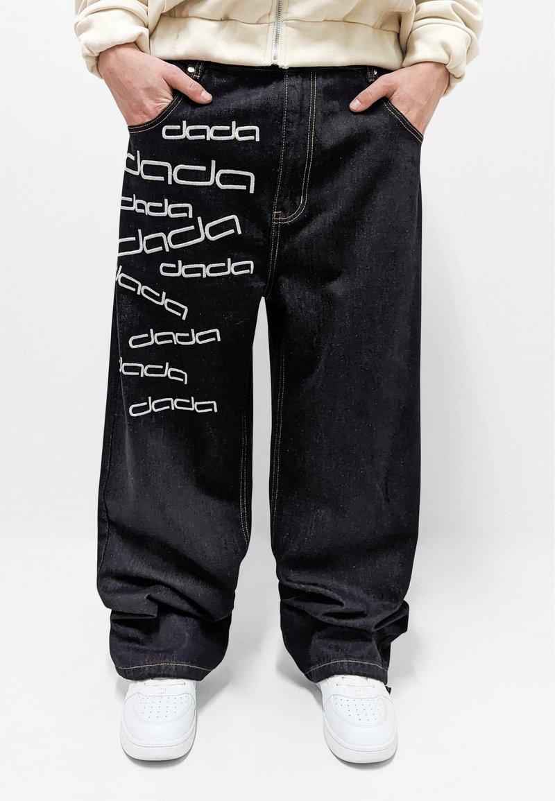 Pantalones de mezclilla negra con corte oversized. Presentan texto blanco "dada" y costuras visibles. Bolsillos frontales y dobladillos con puños, usados con zapatillas blancas.