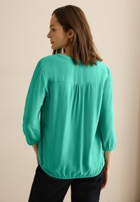 Teal bluse lavet af letvægtsstof, med rund halsudskæring, trekvartærmer og sammensyning i ryggen og forneden.
