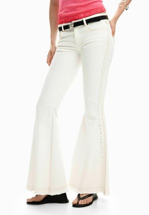 Biele rozšírené džínsy s textúrovaným bočným pruhom, strapcovaným lemom a kontrastným čiernym opaskom. Kombinované s čiernymi žabkami.