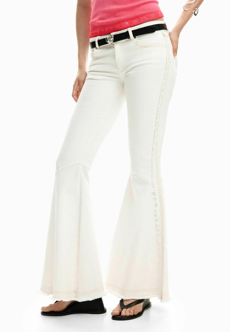 Weiße Schlagjeans mit strukturiertem Seitenstreifen, ausgefranstem Saum und kontrastierendem schwarzem Gürtel. Kombiniert mit schwarzen Flip-Flops.