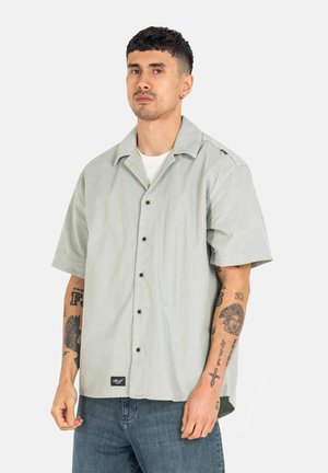 Light green short-sleeve shirt with a collared design, featuring brown buttons and a label near the hem. Worn with blue jeans.