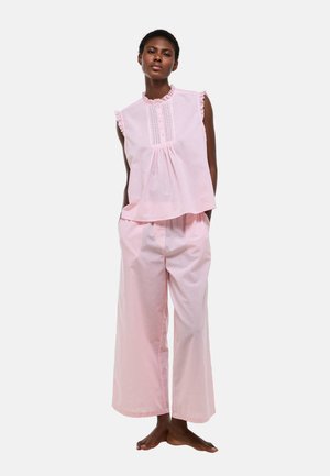 Femme portant un chemisier ample sans manches rose clair avec des volants et un pantalon large assorti, debout pieds nus sur fond blanc.