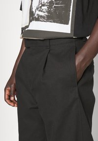 Svarta plisserade byxor med sidofickor, skräddarsydd passform, tillverkade av robust tyg. Hög midja med bältesöglor och subtil textur.