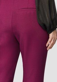 Pantalon ajusté en tissu magenta vibrant, avec une texture lisse, des poches latérales et des manches transparentes noires contrastantes visibles.