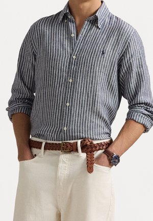 Homme portant une chemise à rayures verticales bleues et blanches boutonnée, manches retroussées, pantalon blanc, ceinture tressée marron et montre-bracelet avec bracelet en cuir marron.