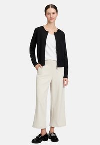 Cardigan noir avec un col rond et des manches longues, présentant des détails de boutons sur les manches, porté sur un haut blanc et un pantalon beige. Chaussures noires.