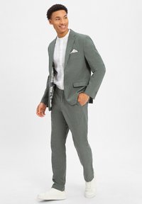 Groen pak van gestructureerde stof, met een enkelvoudig jasje, slanke broek en een wit shirt met een pochet. Witte sneakers.