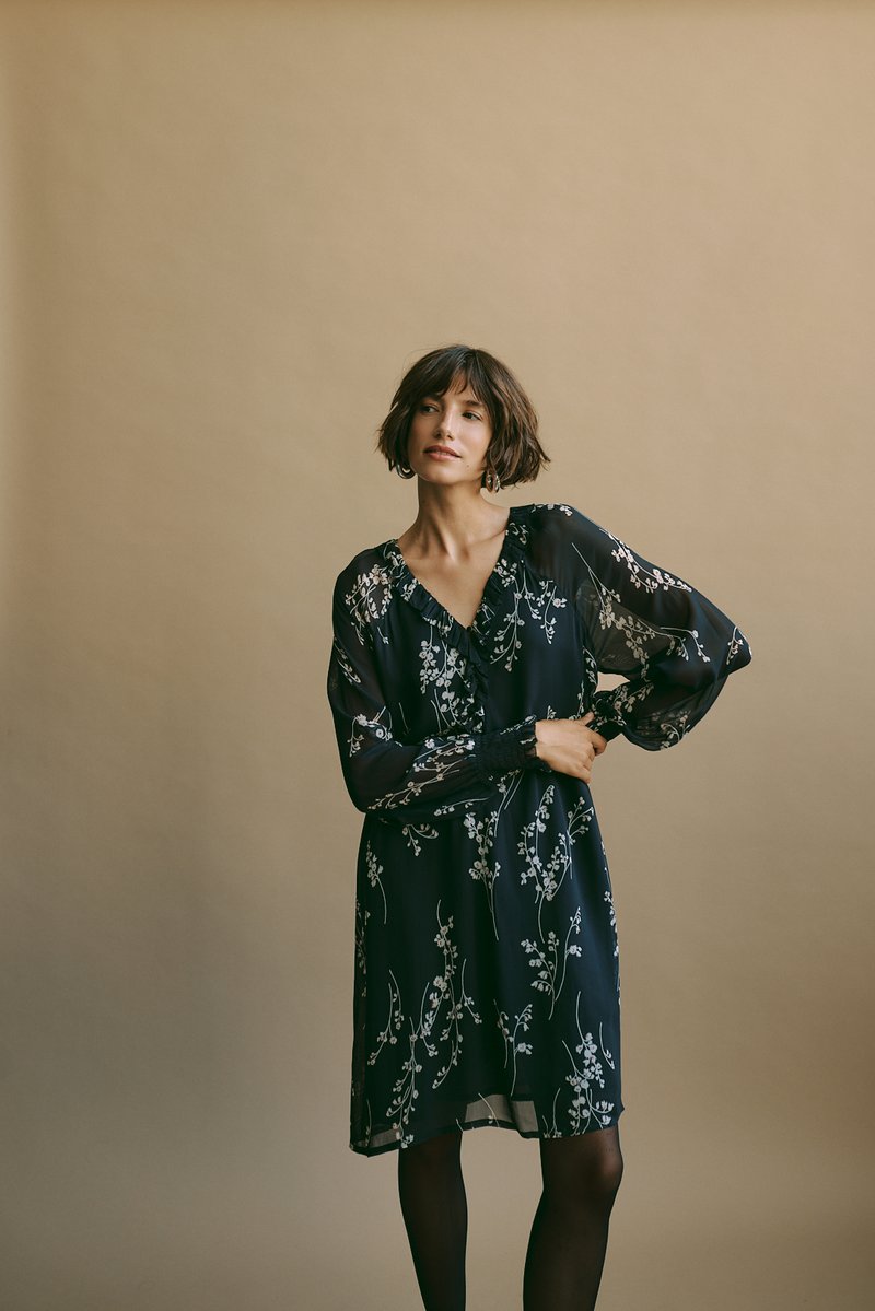 Abito nero a fiori con maniche lunghe e trasparenti e scollatura a V. Presenta una vita aderente e una gonna fluente, lunga fino al ginocchio, con eleganti accenti floreali bianchi.