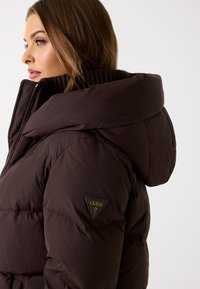 Bruine gewatteerde jas met een grote capuchon, voorzien van een gladde textuur, ritsluiting en een driehoekige logo patch op de linker mouw.