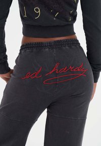 Mörkgråa träningsbyxor med elastisk midja, prydda med röd broderad "ed hardy" text på baksidan, slät textur och figurnära design.