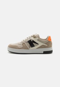 Chaussure de sport beige avec tige en suède et synthétique, logo noir, accent orange au talon et semelle blanche. Bout perforé pour la respirabilité.