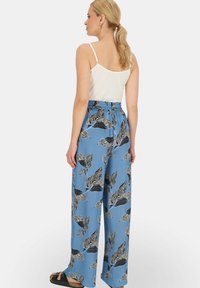 Niebieskie spodnie szerokie z liściastym czarno-białym wzorem, z elastycznym paskiem w talii i miękkiego materiału; modelka ubrana w białą koszulkę na ramiączkach i sandały.