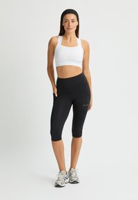 Sostén deportivo blanco con tirantes anchos, combinado con leggins negros hasta la rodilla que cuentan con un bolsillo lateral y detalle de logo. Tejido ligero y diseño ajustado.