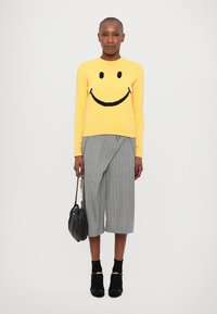 Dzeltena adīta džemperis ar melna smaidoša sejas dizainu, kombinēts ar pelēkiem svītrainiem culottes biksēm. Modelis tur melnu rokassomu; melnas potītes zābaki.