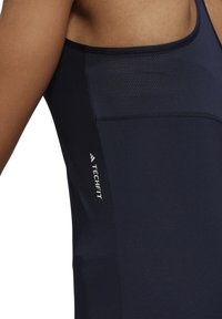 Tmavě modrý sportovní top na ramínka s přiléhavým střihem, hladkou texturou a síťovinou v horní části; obsahuje logo "TECHFIT".