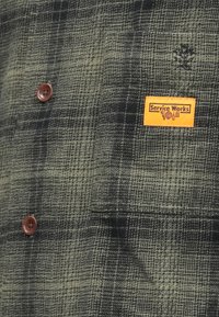 Kurtka zapinana na guziki w ciemnozieloną i czarną kraciastą kratę, z brązowymi guzikami i pomarańczową etykietą "Service Works" na kieszeni na piersi.