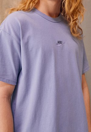 Person med langt krøllet blondt hår har en løs lavendelfarvet Nike T-shirt med et lille logo på venstre bryst.