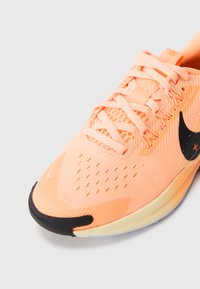 Zapatilla deportiva naranja claro con parte superior de malla, acentos negros en el talón y el swoosh, y detalles texturizados. Los cordones son planos y delgados.