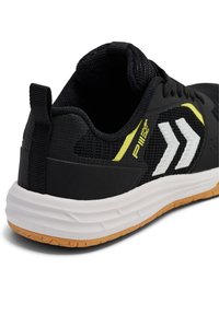 Svart idrottssko med mesh-övre, vita och gula accenter, dragflik och en vadderad vit sula med en strukturerad gummiyttersula.