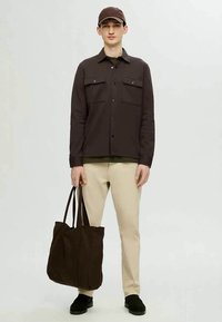 Camisa de botão castanha-escura com dois bolsos no peito, calças de tecido bege e uma bolsa tote castanha-escura. O modelo também usa um boné combinando e sapatos pretos.