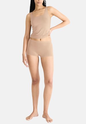 Vrouw staat blootsvoets in een huidskleurige tanktop en boyshort ondergoed tegen een effen witte achtergrond.