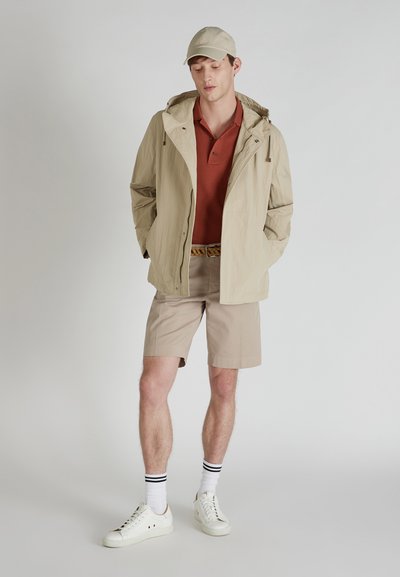Chaqueta ligera beige con capucha sobre una camiseta de polo en color óxido, combinada con pantalones cortos beige, zapatillas blancas y una gorra beige. Vestimenta de manga corta.