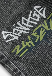 Tissu en denim noir avec des textes style graffiti en blanc et vert olive, ainsi que des éclaboussures de peinture.