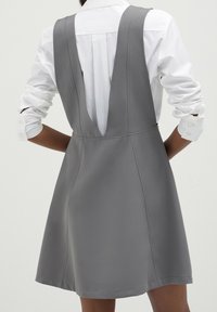 Graues ärmelloses Kleid mit einem tiefen V-Ausschnitt am Rücken, enganliegendem Oberteil, schwingendem Rock und sichtbaren Nähten. getragen über einem weißen Hemd mit Knöpfen.