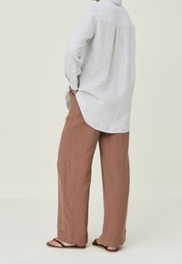 Chemise en lin gris clair à manches longues associée à un pantalon ample en lin marron. La chemise possède un col à boutons et un pli dans le dos.