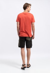 Rode katoenen t-shirt met korte mouwen, gecombineerd met zwarte cargoshorts met groene zakken en accenten, tegen een eenvoudige witte achtergrond.