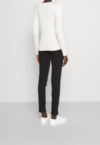 Person som bär en vit kabelstickad tröja, svarta slim fit-byxor och vita sneakers, stående mot en enkel ljusgrå bakgrund.