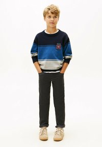 Jongen in een blauwgrijs gestreept sweater, donkere broek en beige sneakers, staand met handen in zijn zakken tegen een witte achtergrond.