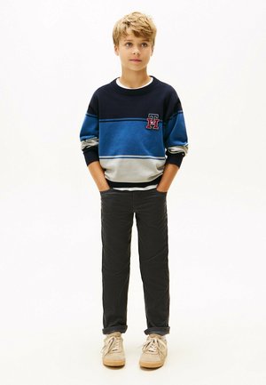 Junge trägt einen blauen und grauen gestreiften Pullover, dunkle Hosen und beigefarbene Sneakers, steht mit den Händen in den Taschen vor einem weißen Hintergrund.